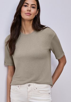 Kobieta o długich ciemnych włosach, ubrana w taupe krótki sweter z krótkim rękawem i białe spodnie, stojąca na tle jasnoszarego jednolitego tła.