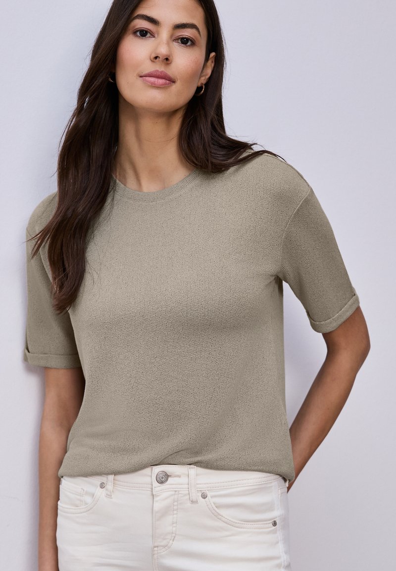 Femme aux longs cheveux foncés portant un haut en maille taupe à manches courtes et un pantalon blanc, debout devant un fond gris clair uni.