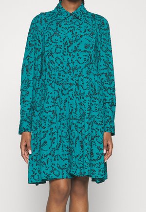 Tealfarbenes langärmeliges Kleid mit schwarzem floralem Rankenmuster, knöpfbarer Vorderseite, Kragen und gestuftem, knielangem Rock, getragen von einer stehenden Person.