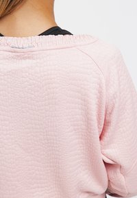 Felpa rosa chiaro con un motivo testurizzato simile a quello di un rettile. Presenta un collo arricciato e una vestibilità rilassata. Sono visibili dettagli delle cuciture in evidenza.
