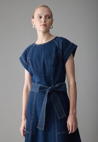 Robe en denim avec des manches courtes retroussées, un col rond, une taille ceinture, et des détails de couture visibles dans une teinte bleu foncé.