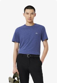 Sininen lyhythihainen t-paita, jossa on pyöreä pääntie, valmistettu pehmeästä kankaasta. Paidassa on pieni valkoinen logo vasemmalla puolella, ja se on yhdistetty mustiin housuihin.