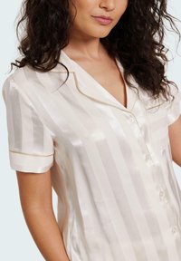 Silk pajama top in cream with vertical stripes and a satin finish. Features short sleeves, a notched collar, gold piping, and button front.