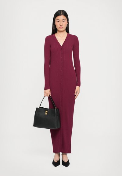 Robe bordeaux longue et moulante à côtes avec un décolleté en V et des boutons sur le devant. Sac à main noir avec quincaillerie dorée et chaussures noires à bout pointu.