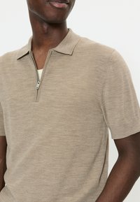 Mann trägt beige Strick-Poloshirt mit kurzen Ärmeln und silbernem Reißverschluss, der teilweise geöffnet ist, vor schlicht weißem Hintergrund.