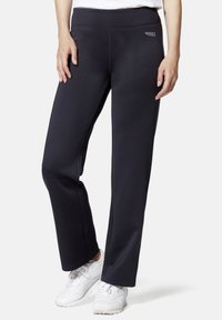 Schwarze Sporthosen mit glatter Textur, hohem Bund und dezentem Logodetail an der Hüfte, kombiniert mit weißen Sneakers.