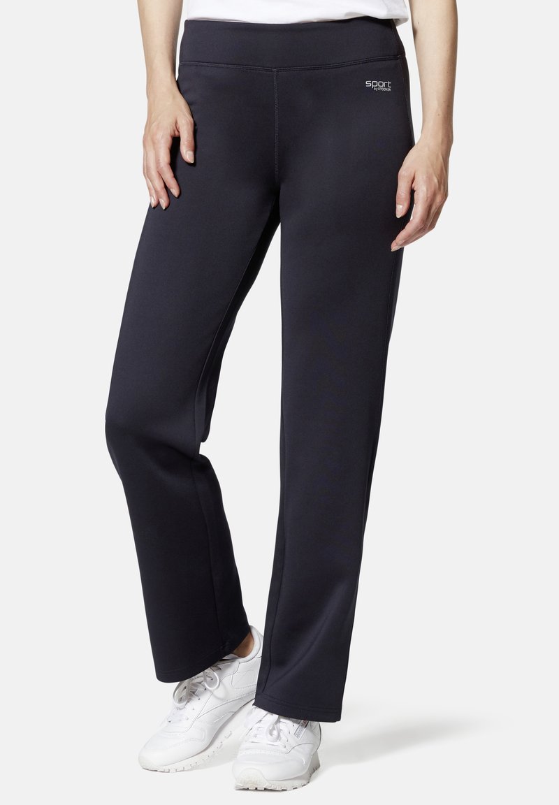 Schwarze Sporthosen mit glatter Textur, hohem Bund und dezentem Logodetail an der Hüfte, kombiniert mit weißen Sneakers.