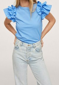 Haut bleu à manches courtes avec des accents volantés sur les épaules, associé à un jean taille haute à délavage clair, doté d'une fermeture classique à boutons et zip.