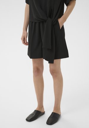 Person iført sort knælange shorts og matchende sorte slip-on sko, stående med den ene hånd i lommen mod hvid baggrund.