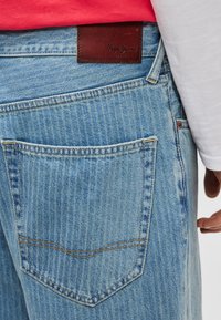 Ανοιχτό μπλε τζιν με κάθετη ριμπ υφή. Διαθέτει καφέ δερμάτινη ετικέτα στη μέση και πίσω τσέπη με κίτρινο ράψιμο.