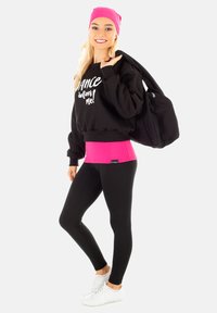 Schwarzes Sweatshirt mit weißen Text, pinkem Bund, schwarze Leggings und weißen Sneakers. Das Model hält eine schwarze Tasche und trägt eine pinke Mütze.