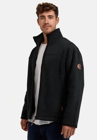Chaqueta de forro polar negra con cuello alto, cierre frontal con cremallera y bolsillos laterales. Presenta un acabado texturizado y un parche de logo en la manga izquierda.
