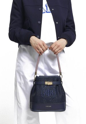 Personne portant une veste marine et un pantalon blanc tenant un sac à main tissé marine avec une anse rayée et un fermoir doré.