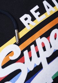 Primo piano di un tessuto nero con il testo bianco "Super" su strisce orizzontali gialle, arancioni, rosse, blu e verdi, e un cordino blu navy con una punta metallica.