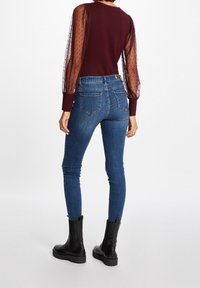 Burgunderfarbenes Top mit durchsichtigen gepunkteten Puffärmeln, kombiniert mit blauen Skinny-Jeans und schwarzen Knöchelschuhen. Glattes Material mit gerippten Bündchen und Nähten.