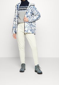 Chaqueta resistente al agua con un patrón de mármol azul y blanco, combinada con un suéter gris y joggers de color crema claro. El calzado son botas de senderismo azules.