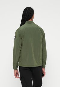 Groene lichtgewicht jas met een gestructureerde afwerking, lange mouwen en een rugpasontwerp. Bevat een patch op de linkermouw.