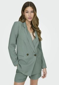 Dubbelbreasted blazer van gedempt groene stof, met twee grote bronzen knopen, en bijpassende shorts. Gladde textuur, geklede pasvorm.