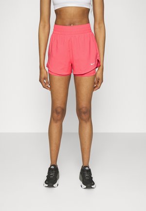 Osoba w koralowo-różowych sportowych krótkich spodenkach Nike, białym krótki topie i czarno-biały obuwiu do biegania, stojąca na jednolitym tle.