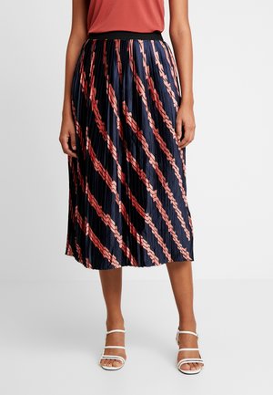 Woman wearing a pleated navy midi skirt with pink and red diagonal patterns, paired with a rust sleeveless top and white strappy sandals.