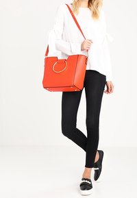 Červená kabelka z umělé kůže se strukturovaným tvarem, zlatými kovovými doplňky a kruhovým držadlem. Nošena s černými legínami a nazouvacími botami s perlovými ozdobami.