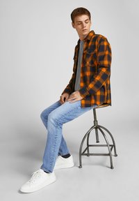 Bărbat tânăr șezând pe un scaun metalic, purtând o cămașă în carouri portocalie și bleumarin, un tricou gri, blugi albastru deschis și adidași albi, privindu-se în jos.