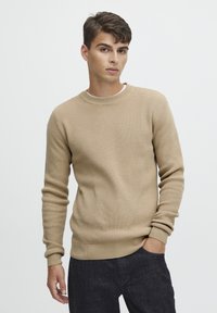 Beige wafelgebreide trui met een ronde halslijn en geribbelde manchetten, gecombineerd met donkere denim broeken. Glad textuur met een aansluitend ontwerp.