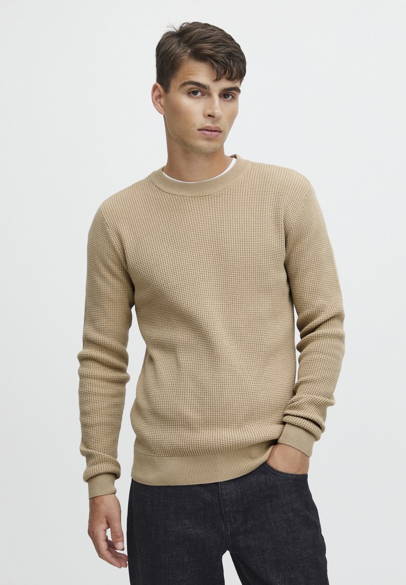 Beige wafelgebreide trui met een ronde halslijn en geribbelde manchetten, gecombineerd met donkere denim broeken. Glad textuur met een aansluitend ontwerp.