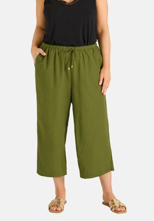 Kvinde iført olivengrønne vidde-bukser med elastisk talje og snøre, kombineret med sort ærmeløs top og beige sandaler.