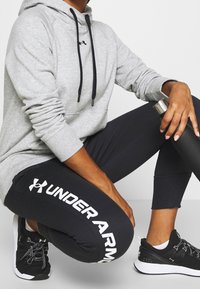 Szara bluza z kapturem z czarnymi sznurkami, czarne legginsy z napisem "UNDER ARMOUR" na białym tle oraz czarne buty sportowe z białymi akcentami.