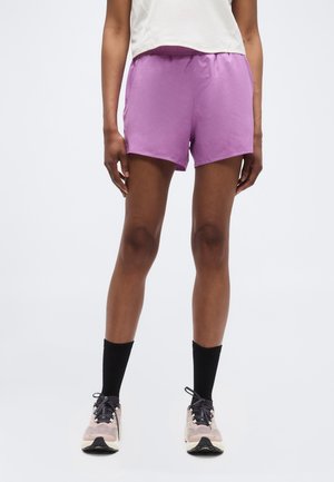 Personne portant un short de sport violet clair, des chaussettes noires montantes et des chaussures de course beiges sur un fond uni.