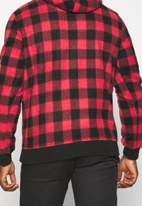 Rot-schwarzes Karomuster Fleece-Hoodie mit Kapuze, gerippten Bündchen und Saum. Der Stoff hat ein weiches, strukturiertes Aussehen und zeigt ein kariertes Muster.