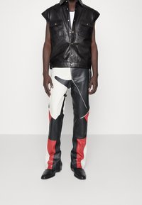 Svart läder utan ärmar med två framfickor, kombinerat med åtsittande byxor i svart, röd och vit läder med abstrakta mönster.