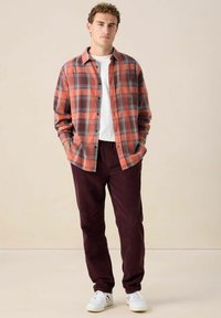 Rood en zwart geruite flanellen shirt boven een wit T-shirt, gecombineerd met bordeaux corduroy broek en witte sneakers. Eenvoudig, casual ontwerp.