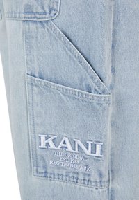 Heledad sinise denimi cargo-püksid, millel on suur küljetasku ja tikkimisega "KANI" logo koos täiendava tekstiga. Pehme tekstuur ja juhuslik disain.