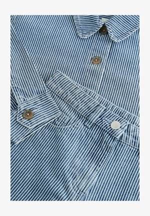 Dryžuotas mėlynas džinsinis marškinėliai su apykakle, turintys rudus mygtukus ir sidabrinį spaudimo mygtuką apatiniame skyriuje. Tekstūruotas audinys, matomos siūlės.