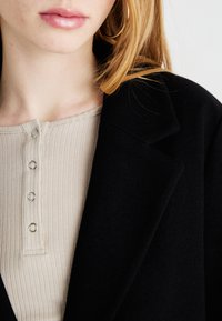 En svart ullkåpe ved siden av en beige ribbestrikket topp med rund hals og sølvfarge detaljer, med fokus på tekstur og fargekontrast.
