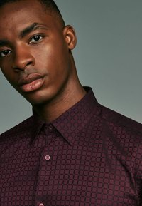 Burgunderfarbene Button-Up-Hemd mit einem Rautenmuster, kurzem Kragen und subtiler Textur. Enthält hellrosa Knöpfe und ein tailliertes Design.