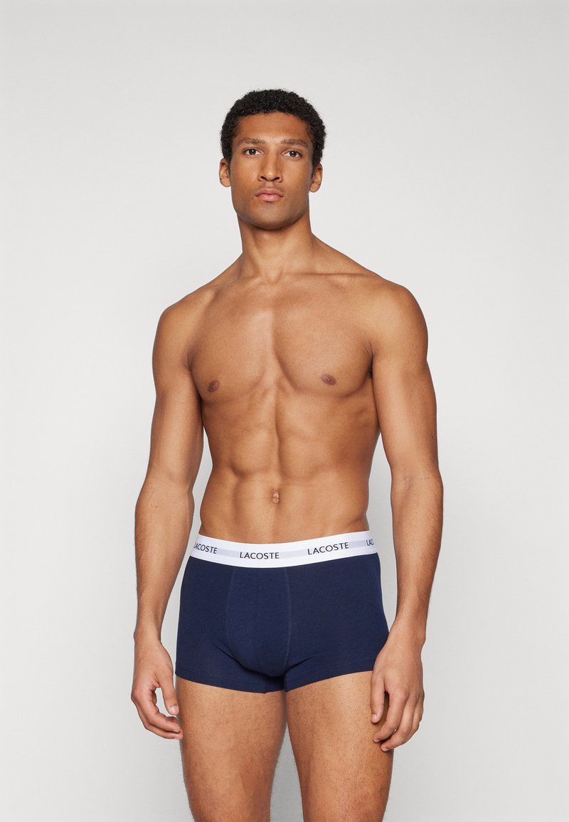 Boxers en coton bleu marine avec une ceinture élastique blanche portant le logo Lacoste. Design ajusté, texture lisse.