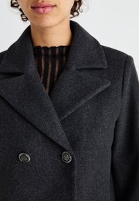 Abrigo de lana gris oscuro de doble botonadura con solapas afiladas y tela negra rayada texturizada visible por debajo. Presenta botones en tono plateado.