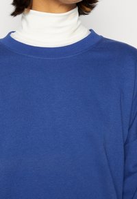 Modrý svetr s kulatým výstřihem, vyrobený z měkké látky. Přes bílý rolák s vrstvami, což ukazuje kontrast barev a textur.