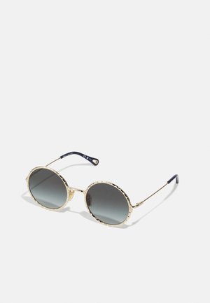 Occhiali da sole oro con lenti rotonde di colore grigio sfumato. Design del bordo texturizzato e bracci metallici sottili con punte blu navy. Eleganti e moderni.