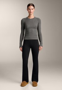 Femeie îmbrăcată cu un top gri închis cu mâneci lungi, cu dungi, pantaloni evazați negri și pantofi slip-on maro, stând în picioare pe un fundal simplu, neutru.