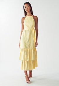 Geel, gelaagd maxi-jurk met halternek en tailleband. De stof lijkt zacht met een gladde textuur, aangevuld met witte sandaaltjes met bandjes.