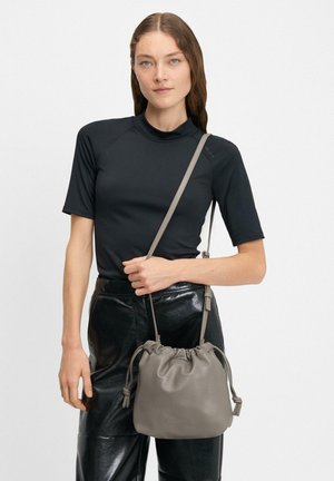 Bolso de cubo de cuero gris con cierre de cuerda, textura suave y correa ajustable, sostenido por una persona que lleva una blusa negra ajustada y pantalones brillantes.