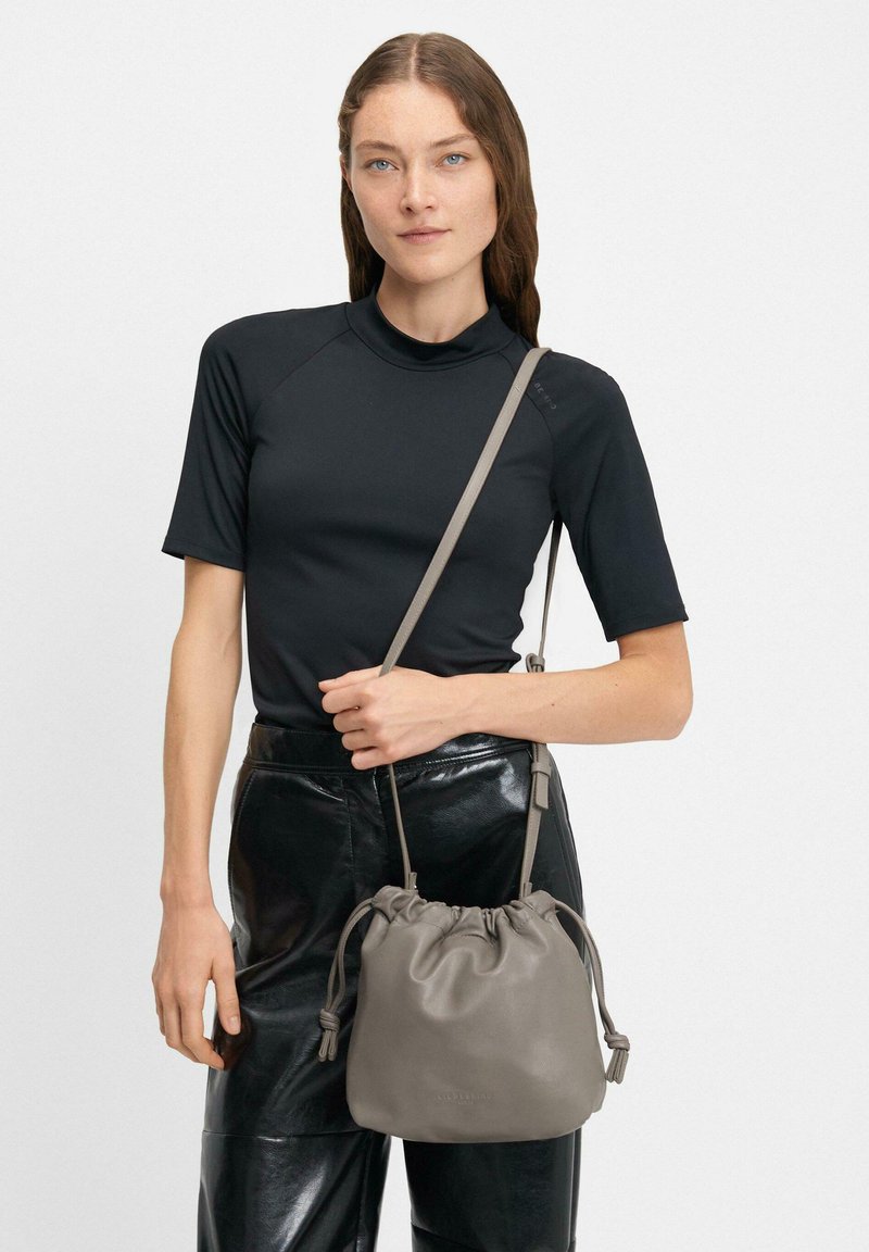 Borsa a secchiello grigia in pelle con chiusura a coulisse, texture liscia e tracolla regolabile, tenuta da una persona che indossa un top nero aderente e pantaloni lucidi.