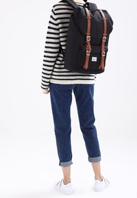 Musta reppu ruskeilla nahkahihnoilla ja soljilla, valmistettu kestävästä materiaalista; henkilöllä on päällä raidallinen neuletakki ja käärityt farkut.