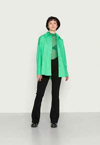 Camisa oversized verde com fecho de botão, usada sobre um top verde estampado, combinada com calças pretas de boca de sino e sapatos pretos, em um ambiente minimalista.