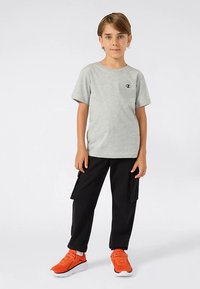 Zēns stāv uz priekšu vērsts, ģērbts pelēkā t-kreklā, melnās biksēs-kargo un spilgti oranžās tenisa kurpēs uz vienkārša balta fona.