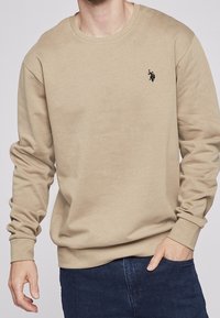Uomo che indossa un maglione beige a maniche lunghe con collo rotondo, dotato di un piccolo logo ricamato nero sul lato sinistro del petto, abbinato a jeans blu scuro.
